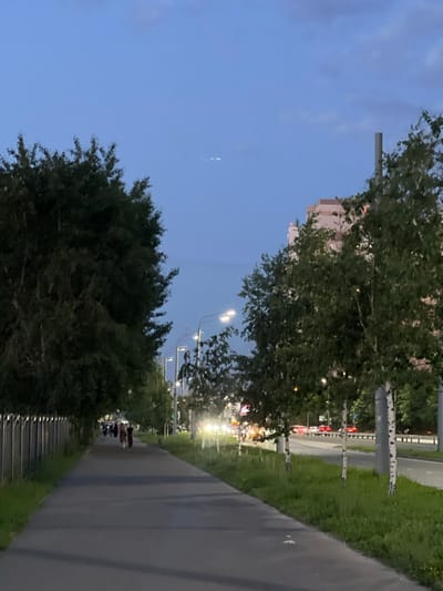 The image depicts a twilight scene along a paved walkway adjacent to a road. On the left, a tall, dark green dense tree line runs parallel to a metal fence. Several figures, appearing to be people, are walking away from the camera on the walkway. To the right of the walkway, a strip of green grass is visible, followed by a multi-lane road with moving vehicles indicated by headlights and taillights. Several streetlights are illuminated, casting light on the scene. In the background, a tall, light-colored building is visible, and a grey cylindrical structure stands near it. The sky is a deep blue with a few faint clouds, and three small white lights appear as a formation in the upper portion of the sky. This scene is located in Vysokaya Gora, Russia.