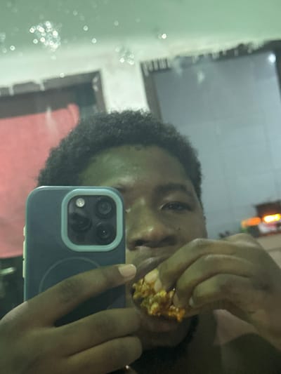 The image captures a reflected self-portrait of a dark-skinned individual with short, dark, curly hair. The subject holds a dark blue smartphone, featuring a triple-lens camera array and flash module within a light blue rectangular bezel, in their left hand, positioning it for the photograph. Simultaneously, their right hand grasps a piece of food, characterized by a textured, reddish-orange exterior, which is positioned at the subject's mouth. The background, visible as a reflection, contains scattered out-of-focus light sources across the upper portion. To the left, a blurred reddish vertical surface is present, while to the right, a darker vertical surface is observed. Further to the lower right, an indistinct object emitting orange and yellow light is reflected. The scene occurs in Ipetumodu, Nigeria, under ambient light conditions suggesting evening or night.