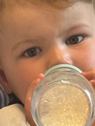 A close-up shot depicts a baby drinking from a clear bottle. The baby's face, with light skin and blue eyes, is the main focus. The bottle, filled with a clear liquid, is positioned against the baby's face, obscuring part of it. A hand is holding the bottle. The baby is wearing a blue and white striped garment with a white cloth nearby. The image is taken at a close, tilted angle. The location and city are not discernible from the image.