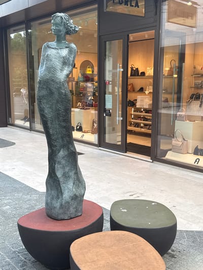 This is a vertical, eye-level, outdoor shot of a bronze statue of a woman in front of a storefront. The statue depicts a woman from the chest up, with her head tilted slightly down and her hair styled in an updo. Her arms are not visible. The statue has a textured, weathered appearance.
Behind the statue is a storefront with large glass windows, revealing the interior of a boutique. The store appears to be selling handbags and possibly shoes, with various items displayed on shelves and mannequins. The interior is brightly lit and has wooden accents. The word "FURLA" is visible on a sign on the left side of the storefront.
To the right of the statue, there are three large, abstract, circular sculptures. The topmost is a pale green, the middle one is a light brown, and the bottom one is dark red. These sculptures are placed on a textured, light-colored paved surface. The ground in front of the storefront is a mix of grey cobblestones and lighter paving stones.
The lighting suggests it is daytime, possibly late afternoon or early evening, as there are some shadows, but the scene is well-lit. The weather appears to be clear. The presence of a retail store and outdoor sculptures suggests an urban or commercial setting. There are no people clearly visible in the statue or the storefront, though reflections in the window might obscure some details.