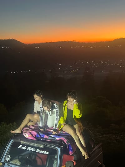Three young women are seated on the roof of an open-top vehicle at night. The vehicle is a light blue Jeep-style car with a sticker on the windshield that reads "Roda Batur Sunrise." A colorful blanket is spread on the roof, providing a seating surface for the women. The woman on the left wears a white shirt and shorts, with her legs bare. The woman in the middle wears a white jacket with black stripes and black shorts. The woman on the right wears a neon yellow jacket over a black top and shorts. The background depicts a panoramic view of a city at night, with numerous lights twinkling in the distance, nestled among hills. The sky above is transitioning from dusk to night, with the last remnants of a bright orange sunset visible on the horizon, transitioning to a deep blue. The location is identified as Kintamani, Indonesia.