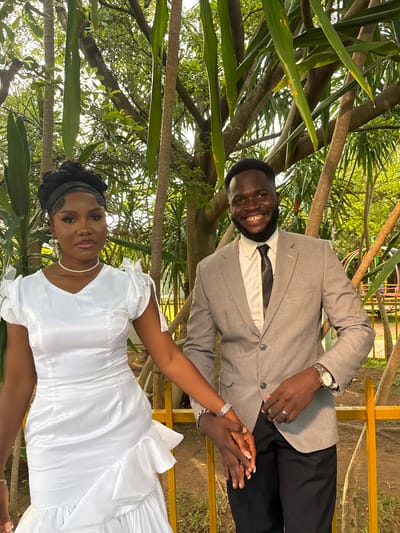 A wedding photograph is depicted in the city of Barikin Gidar Kanat, Nigeria. A Black couple, likely the bride and groom, stand outdoors, holding hands. The bride is on the left, wearing a white, knee-length dress with ruffled detailing at the hem and puffed sleeves. She has her hair styled in an updo and is adorned with a pearl necklace and dangling earrings. The groom stands to her right, dressed in a light grey suit jacket, a white shirt, and a black tie. He has a beard and is smiling at the camera. They are positioned in front of a backdrop of lush greenery, with trees and foliage surrounding them, suggesting a garden or park setting. A yellow railing is visible in the foreground.