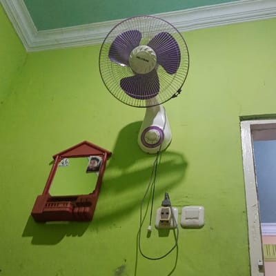 This image captures an interior scene in Ketowan, Indonesia, focusing on a lime green wall. A white wall-mounted fan with purple blades and a metallic cage, branded "GELIJIANG", is positioned in the upper-center. Its power cord extends downwards. To the left and below the fan, a rectangular mirror is housed within a reddish-brown, house-shaped plastic frame attached to the wall. The mirror's surface displays a green sticker with partially legible text and a small photograph of a male individual. Below the fan, a white electrical outlet is mounted, with a black power cord plugged into it. To the right of the outlet, a white single-toggle light switch is present. The lime green wall exhibits minor scuff marks and faint cracks. A lighter green ceiling with white crown molding is visible at the top. On the far right, a partial view of a doorway with a white frame reveals a glimpse of an adjacent room featuring a light blue wall and white crown molding. Shadows from the fan and mirror are cast upon the wall.