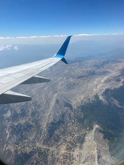 An aerial view from an airplane window shows a mountainous landscape under a clear blue sky with scattered clouds. The wing of the airplane is prominently visible in the foreground, with its white surface and blue wingtip extending into the frame from the left. The terrain below is rugged and varied, featuring rocky outcrops, sparse vegetation with scattered trees, and areas covered in dense green forest. The topography suggests a significant elevation change, with valleys and slopes visible. Faint traces of roads or paths can be seen winding through the mountains. In the distance, layers of mountains fade into a hazy blue, indicating atmospheric perspective. The scene is devoid of any visible human presence or structures, suggesting a remote, natural environment. This perspective is consistent with flying over the mountainous region surrounding Bucak, Turkey, known for its rugged terrain.