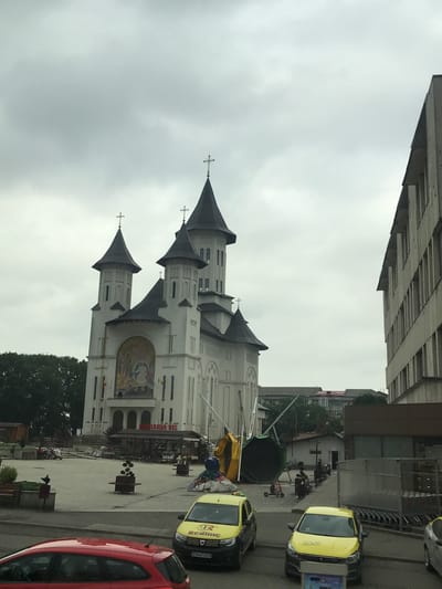 This image captures a wide-angle view of an urban square in Fălticeni, Romania, under an overcast sky.
The prominent structure on the left is a large, white church with five dark, conical spires, each topped with a cross. The central spire is the tallest. The church facade features a large, colorful mosaic or mural above a series of arched windows and entrances. Below this mural, a sign reads "LIBRĂRIA VIS". Two small flags, consistent with the Romanian national flag (red, yellow, blue vertical stripes), are mounted on the facade. To the far left of the church, a dense canopy of green trees is visible.
On the right side of the image, a multi-story, light-colored building with numerous vertical window recesses stands prominently, extending beyond the frame. Its architecture appears utilitarian.
The foreground and midground consist of a paved public square. A street runs across the immediate foreground, where a red hatchback car is partially visible on the left. Two yellow taxi cabs, both with "TAXI" branding and one additionally marked "REDLINE," are parked or driving on this street. The license plate "SV-47 VER" is partially visible on one of the taxis.
Within the paved square, several dark-roofed market stalls or kiosks are arranged in front of the church. Various planters containing small trees or shrubs are distributed throughout the square, along with benches. A pigeon is perched on a planter or bench in the lower-left midground.
Towards the right side of the square, a large, partially collapsed or disassembled structure, appearing cylindrical and made of yellow and dark green/black material, is visible. It is supported by poles or scaffolding. Near this structure, two individuals are seen operating electric scooters. In the far right foreground, adjacent to the multi-story building, a metal rack holding numerous shopping trolleys or carts is present.
Several people are scattered throughout the square, walking or standing near the stalls and benches. The sky is uniformly gray and overcast, providing diffused lighting to the scene. In the distant background, various residential or smaller commercial buildings with different rooflines are discernible.