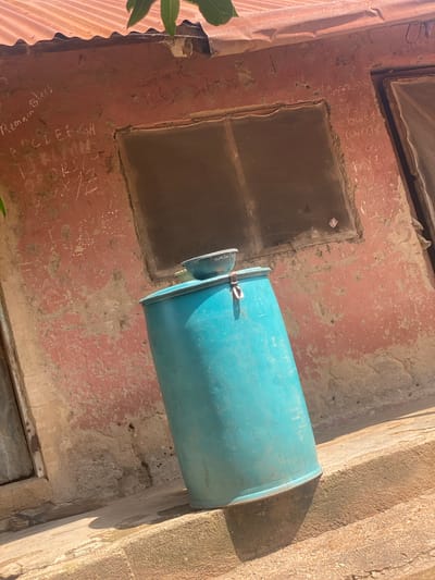 The image depicts a weathered, reddish-pink building facade with graffiti scrawled in white chalk. The graffiti includes what appears to be an alphabet sequence and some unintelligible text. A blue plastic barrel with a small bowl on top stands tilted on a concrete ledge in front of the building. A corrugated metal roof is visible at the top of the frame, with some greenery peeking from the left side. The scene appears to be outdoors, possibly in a semi-urban or rural setting, given the rough texture of the walls and the simplistic construction suggested by the roof. There are no people visible in the image. The overall impression is one of decay and rudimentary living conditions. The location is identified as Daniel, Nigeria.