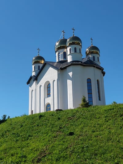 The image shows a white Orthodox church with golden domes situated on a grassy hill under a clear blue sky. The church features multiple tiered domes, each topped with a cross. The building has arched windows and a dark-colored roof. A few small evergreen trees are visible on the hill near the church. The environment is natural, with a focus on the architectural structure of the church against the backdrop of the sky and vegetation. The image does not contain any people or depict any actions or interactions. The location is identified as the city of Zashchita, Russia.