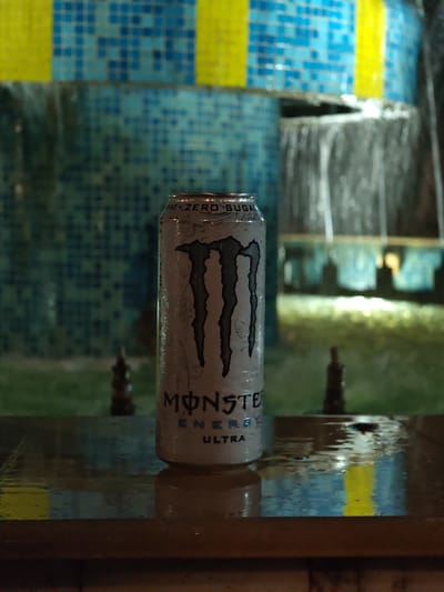 A single, cold Monster Energy Ultra "Zero Sugar" can stands upright on a dark, wet, reflective surface in the foreground. Condensation and water droplets are visible across its white and black metallic body. The surface below reflects the can and elements of the background, also showing numerous water droplets. In the soft-focused background, a tiled water feature or fountain is visible. The tiles are predominantly small squares in various shades of blue, interspersed with sections of yellow tiling. Water cascades downward from a ledge within the structure. The scene is under low, artificial illumination, creating reflections and a distinct atmosphere. No people or other subjects are present. The image is stated to be located in Veliki Preslav, Bulgaria; however, no identifiable landmarks or unique features within the image confirm this specific geographical location.