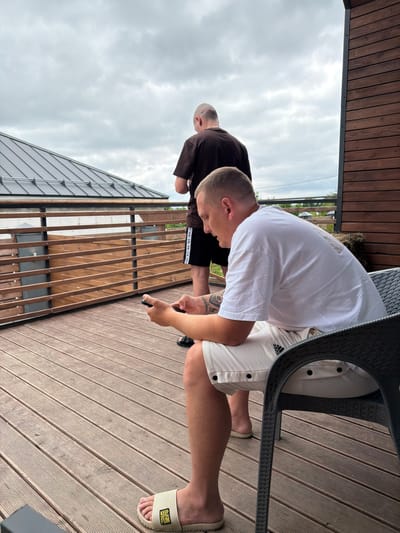 Two men are on a wooden deck. One man is standing and looking out. The other man is sitting in a chair and looking at his phone. The deck has a wooden railing and a wooden roof. The sky is cloudy. The scene is in the city of Semiluki, Russia.