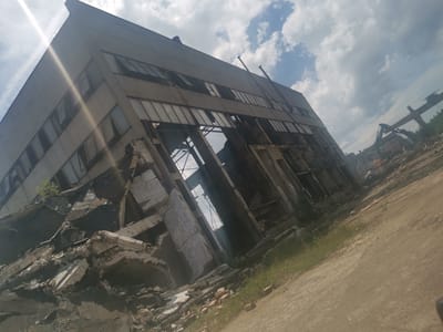 The image captures a desolate industrial scene in Zhivkovo, Bulgaria, dominated by a large, partially demolished concrete building under a partly cloudy sky. The time of day is clearly daytime, evidenced by the bright, diffused sunlight and a prominent sunbeam or lens flare emanating from the upper left of the frame, casting a bright streak across the structure.
The main subject is a multi-story, grey concrete industrial building, likely a factory or warehouse, showing extensive damage. The left side of the building, particularly its lower sections, has largely collapsed, leaving a substantial pile of concrete rubble, fractured beams, and exposed rebar at its base. Many windows throughout the building are broken, missing, or boarded up, and sections of the wall have given way, revealing the interior structural framework and allowing views through to the sky.
The ground in the foreground and around the base of the building is unpaved, consisting of dirt and gravel, with patches of sparse green grass and scattered red bricks from the debris. In the mid-distance and background, more signs of industrial activity or demolition are visible. To the right, heavy machinery, possibly an excavator or part of a crane, stands amidst further rubble and derelict structures, suggesting ongoing demolition or a long-abandoned site. The overall impression is one of decay, destruction, and an industrial landscape in a state of disuse or transformation. No people are visible in the image, and no discernible text can be seen.