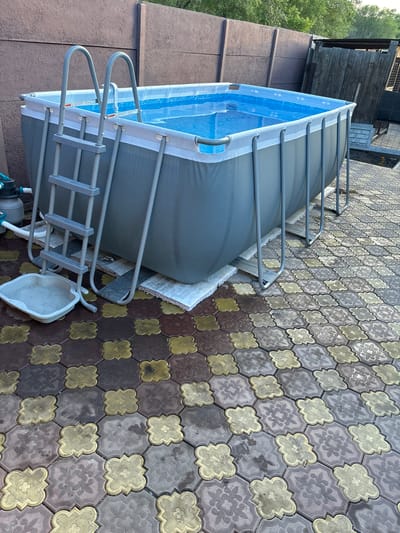 An above-ground swimming pool is set up on a patterned patio. The pool is rectangular with a grey liner and a metal frame, filled with blue water and equipped with a ladder. A white foot bath is placed on the patio in front of the ladder. The patio is paved with interlocking concrete blocks in shades of brown, grey, and yellow. In the background, a brown fence separates the yard from trees and vegetation. A small wooden structure, possibly a shed or outhouse, is visible to the right of the pool. The image depicts an outdoor residential setting.