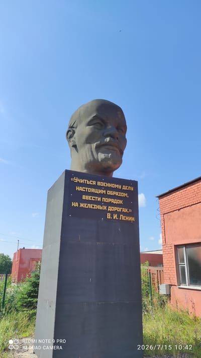 A bust of Vladimir Lenin stands on a tall, dark pedestal against a clear blue sky. Golden text is inscribed on the pedestal. To the right of the pedestal is a red brick building with a window. Green grass and trees are visible in the foreground and background. The image also includes a timestamp in the bottom right corner: "2026/7/15 10:42" and a watermark in the bottom left corner: "REDMI NOTE 9 PRO AI QUAD CAMERA". The bust is facing towards the left of the frame.