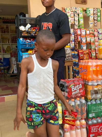 An outdoor market scene presents two people. An adult with a black t-shirt stands near a boy wearing a white tank top and colorful cartoon-printed shorts. The boy holds a bag of "McFries." The market behind them is stocked with various items: beverages like "Squeeze Orange" and "Beta Malt," alongside packaged snacks and drinks. The merchandise is neatly arranged on shelves, creating a vibrant backdrop. The scene suggests a casual moment captured amidst a local commercial setting.