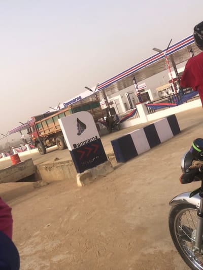 A road scene features a motorcycle with its front wheel and part of the handlebars visible on the right. A sign reading "Danmarna" with a logo and arrows is placed near a striped pillar with alternating white and navy blue bands. A truck sits alongside the sign. The backdrop includes buildings decorated with banners that have red, white, and blue stripes. The sky appears pale.