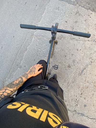 An aerial, high-angle shot shows a person standing over a black bicycle, presumably in Luis Guillón, Argentina. The person's tattooed arm is visible, resting on their leg. The bicycle is black, with black handlebars and a black pedal visible. The person wears a black t-shirt with yellow lettering. The ground appears to be concrete, with cracks and variations in color.