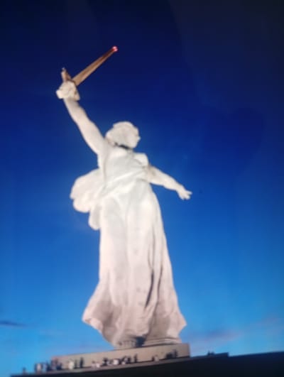 A low-angle, slightly blurry, full shot shows a large white statue of a woman holding a sword above her head against a bright blue sky. The statue, which appears to be made of concrete or stone, is illuminated from below, creating highlights and shadows. The woman is depicted with her right arm raised, and the sword is angled upwards, with a bright red object at its tip, possibly a light or a flame. Her left arm is bent at the elbow, with her hand extended forward. She is wearing a long, flowing robe. At the base of the statue, a dark, out-of-focus structure is visible, with several small, indistinct figures standing on it. The overall scene suggests a monument or memorial. This monument is the Motherland Calls statue located in Mamayev Kurgan, Volgograd, Russia, not Orenburg.