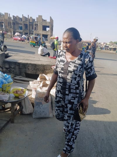 An outdoor scene in Kwekwe, Zimbabwe, depicts a bustling street environment under clear daylight. In the foreground, a woman of dark complexion, with dark hair pulled back, is shown mid-stride, walking towards the viewer's right. She wears a black and white geometrically patterned jumpsuit with a V-neckline and carries a small, leopard-print shoulder bag on her right side, suspended from her left shoulder. White footwear is visible on her left foot. Her gaze is directed slightly downwards and to her left.
Immediately to her left, a small, round table on a wooden stand holds a yellow plastic bowl containing several brown eggs, a yellow woven basket filled with blue material (possibly plastic bags or fabric), and a clear plastic bag. On the ground adjacent to the table, several beige and white sacks are stacked, one clearly labeled "SUPAS" with an elephant logo. These items are situated on a concrete surface, bordered by a raised concrete curb or platform that runs across the mid-ground.
Beyond this platform, a paved road is visible. On the road, a green and yellow auto-rickshaw (tuk-tuk) is present. Further down the street, a red car is parked or moving. Numerous individuals are scattered throughout the background: one person in a white shirt and dark pants is squatting or bending over near the auto-rickshaw, while another in a bright green shirt with a dark backpack stands nearby. More people are observed further in the distance, some under umbrellas, suggesting market activities. The distant background on the left is dominated by a large, multi-story concrete building that appears unfinished or abandoned, characterized by exposed concrete, multiple empty window frames, and structural elements. To the far right in the distance, additional market stalls or informal structures are visible. The ground throughout the scene appears to be a mix of concrete and dusty earth.