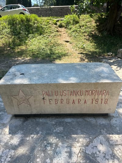 In Škaljari, Montenegro, a concrete memorial stands in a sun-dappled outdoor setting. The rectangular monument is engraved with red lettering and features a red hammer and sickle symbol. The inscription reads "PALI U USTANKU MORNARA FEBRUARA 1918," commemorating those who fell in the uprising of sailors in February 1918. The memorial is placed on a stone-paved area, with a lush green hillside rising behind it. A white car is visible on the hillside, and a large tree casts shadows over the scene. The environment appears to be a historical or commemorative site, likely in a park or public space.