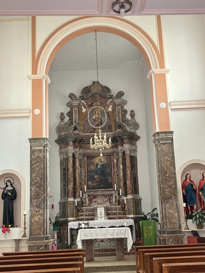The image shows the interior of a church in Taormina, Italy, likely a cathedral or a large parish church, with an altar and religious statues. The altar is adorned with a white tablecloth and is surrounded by candlesticks and religious iconography. To the left of the altar, there is a statue of a robed female figure holding a cross, and to the right, there are two statues of female figures in robes. The church features ornate architectural elements, including marble columns and decorative arches. The overall atmosphere is solemn and reverent, typical of a place of worship.