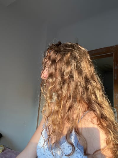 The image shows a person with long, wavy, golden-brown hair, wearing a light blue floral patterned top with lace trim. The person is positioned so their head is angled downward towards the left side of the image. The background is a neutral wall. There's a window frame visible in the background. No information within the image confirms the location is in Saarbrücken, Germany.