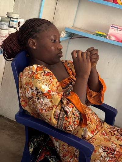 An indoor, eye-level shot taken in Ikot Ekpene, Nigeria, features a woman seated on a blue plastic chair. She has dark skin and reddish-brown braided hair, styled in cornrows with a bun at the back. She wears a vibrant patterned dress or top in shades of yellow, orange, and red, with a plain orange layer visible beneath. Her hands are clasped together in front of her, and she looks upwards and slightly to her left with a neutral, observant expression. A small stud earring is visible in her left ear.

Behind her, to the left, is a light-colored shelf or table with a blue trim. On this shelf, a prominent pink and white box displays the text "Breclomot" with "Aceclofenac & Paracetamol 100 + 500" written below it. Several white plastic containers are also visible; one clearly shows "CALCITONE" on its label, and another has partially visible text resembling "BSULI". The background consists of a light-colored wall and a neutral-toned floor, suggesting a simple, functional space, likely a waiting area within a clinic or pharmacy. The lighting indicates it is daytime.