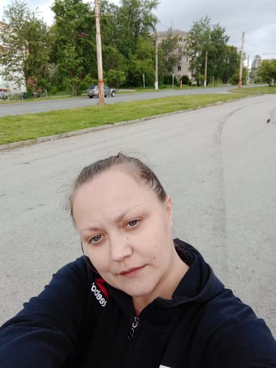 The image captures a female individual taking a selfie outdoors in a residential area of Berezniki, Russia, under an overcast sky during daylight hours. The individual, appearing to be in her 20s to 40s, has light skin, light-colored eyes (greenish-blue), and brown hair pulled back, with visible flyaways. She is wearing a black zippered hoodie or jacket, with a reversed "Reebok" logo visible in white and red on her left chest area. Her expression is neutral as she looks directly into the camera.

The background shows a grey asphalt road with visible tire marks and stains, bordered by a concrete curb and a strip of vibrant green grass on the left. Further back, another section of the asphalt road curves to the right. A silver or grey compact SUV is visible on the far left, moving away from the viewer. Multiple utility poles, some wooden and some concrete, with overhead wires, line the road. Lush green deciduous trees are abundant, suggesting a summer or late spring season. Behind the trees on the left, a light-colored building is partially visible. On the right side of the frame, in the distance, a multi-story apartment building with numerous windows and a grey facade, consistent with Soviet-era architecture, stands behind the trees. A small, dark-feathered bird (likely a pigeon or sparrow) is visible on the asphalt road to the far right. The overall scene depicts a typical urban street in a residential district of Berezniki.