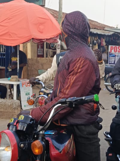In Kabong, Nigeria, a street scene unfolds near a market stall. An individual wearing a hooded, patterned jacket sits on a red motorcycle, facing away from the camera. Their left hand rests on the motorcycle's handlebars, and their right arm is bent, partially obscuring their face. The motorcycle has a prominent headlight, orange turn signals, and the word "BOXER" visible on its side.
Behind the motorcycle rider, another person in a light-colored shirt is visible. To the left, a person in a dark shirt is standing by a white table laden with stacks of cash and what appears to be promotional flyers. One flyer on the table advertises "POS Service Available HERE" and shows images of a phone and a POS machine, along with a phone number and a green circular logo.
A large, bright orange umbrella provides shade over the area. In the background, market stalls with corrugated metal roofs are visible, displaying various goods. The ground is paved, and the overall impression is of a busy, informal commercial setting.