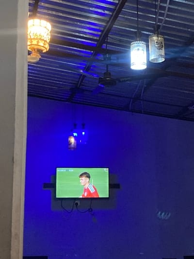 The image depicts an interior scene, likely a room or bar, with a television displaying a soccer match. The room is illuminated by blue lighting and features a corrugated metal ceiling with exposed beams and a ceiling fan. Hanging from the ceiling are three decorative lanterns, one with an ornate, golden design and two cylindrical white lanterns with patterned detailing. A white pillar is visible in the foreground on the left side of the frame. The television, mounted on the wall, shows a young man in a red soccer jersey with white stripes on the sleeves. The overall ambiance is dimly lit, with the blue lights creating a vibrant and somewhat atmospheric setting. This scene is located in Gwarinpa, Nigeria.