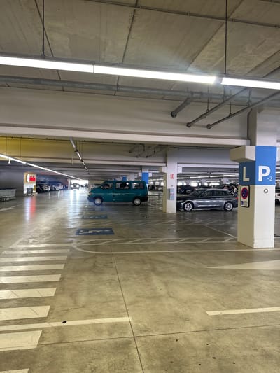 This is a photograph taken in a multi-story parking garage, likely in Alfafar, Spain, as indicated by the location context. The perspective is from above, looking down into the garage. The scene is illuminated by long fluorescent lights running parallel to the rows of parking spaces.
The primary subjects are cars parked in their designated spots. A grey sedan is visible in the upper portion of the image, parked under a blue and white sign that reads "P L2". A teal-colored van is parked further down the row, and several other vehicles are visible receding into the distance. The floor of the garage is concrete, marked with white lines indicating parking bays and directional arrows. There are also blue symbols on the floor indicating disabled parking spaces.
The setting is an indoor parking facility, characterized by concrete pillars, overhead lighting, and a general utilitarian aesthetic. There are no visible people in the frame, suggesting it might be a quiet time. The time of day cannot be determined from the image, and the weather is irrelevant as it is an indoor setting.
Notable details include the signs indicating parking levels and accessibility, as well as the reflective surface of the floor, likely from a polished concrete finish or a light coating. There are also visible pipes and conduits running along the ceiling and walls, typical of infrastructure in such a building. A red and white sign with text, possibly a brand or advertisement, is visible further down the row. The overall impression is one of order and functionality.