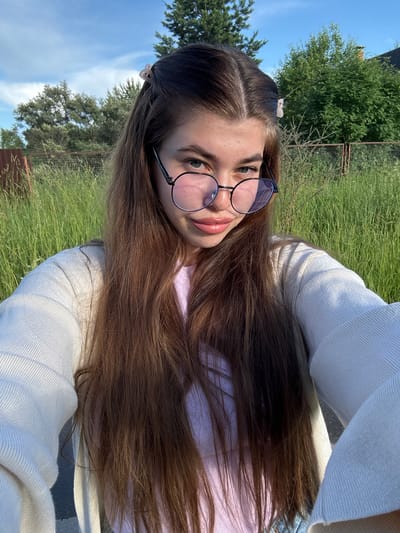 A young woman with long, brown hair and glasses is taking a selfie outdoors. She is wearing a light-colored sweater over a pink top. Her hair is partially visible, framing her face and falling down her chest. She has light-colored eyes and her lips are slightly parted. The background consists of tall green grass and trees, with a blue sky and some clouds visible. There is also a hint of a fence in the background. The lighting suggests it is daytime, possibly late afternoon given the warm tones. The overall scene appears casual and relaxed. The image seems to have been taken in a rural or park-like setting. There is no visible text in the image.