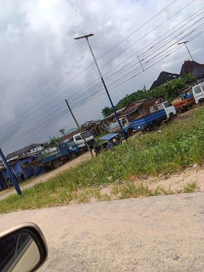 The image shows an outdoor scene with a cloudy sky and power lines crisscrossing the frame. In the foreground, a portion of a car's side mirror is visible, reflecting a purple object. The midground features a dirt road with several parked trucks. One blue truck is prominently displayed, with other white and blue trucks behind it. Some structures, appearing to be makeshift shelters or buildings with corrugated metal roofs and blue tarps, are visible behind the trucks. Lush green grass and vegetation grow alongside the road. In the background, houses and trees are visible, with a distinctive building with a pointed roof. The scene appears to be an informal market or a depot for trucks, likely located in Yenegoa, Nigeria, based on the context provided. There are no people actively engaged in the scene.