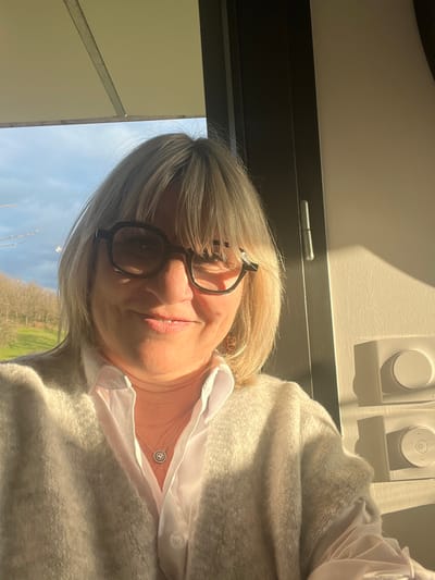 A blonde woman with bangs and dark-rimmed glasses smiles directly at the camera in what appears to be a selfie, located in Chambœuf, France. She wears a white collared shirt and a light-colored, possibly off-white, cardigan. A silver necklace with a circular pendant rests on her chest. Strong sunlight from the right illuminates her face and the interior space. To her left, a window reveals a clear blue sky with scattered clouds above a landscape of bare trees and green terrain. To her right, a light-colored interior wall features two white, rectangular wall-mounted devices, each with a circular control, casting shadows due to the direct sunlight. A dark vertical frame element, possibly part of a window or door, with a visible hinge, is situated between the woman and the wall devices.