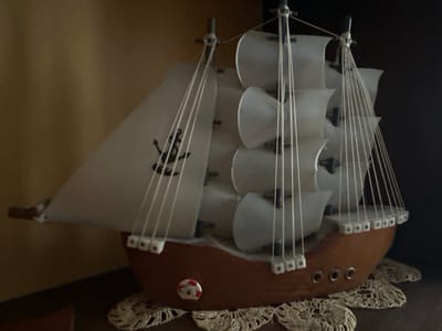 A model sailboat sits on a doily. The boat has a brown hull, white sails with a black anchor design on one, and white ropes. It is situated indoors, with a brown wall visible on the left and a dark surface on the right. The location is unspecified; the city of Troyan, Bulgaria is also not visible in the image.