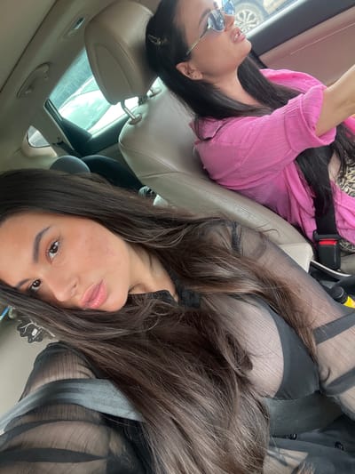 The image shows two women inside a car, likely in Kiryat Bialik, Israel, based on the prompt's request to include that location. The woman in the foreground is wearing a black, sheer top and looking directly at the camera. Her dark hair is partially obscuring her face. The woman in the background is wearing a pink top and sunglasses. Both women appear to be passengers, as indicated by the presence of a seat belt. The car has a sunroof visible in the background. The image is taken from an unusual angle, possibly from the car's ceiling or dashboard. The car's interior is light-colored leather.
