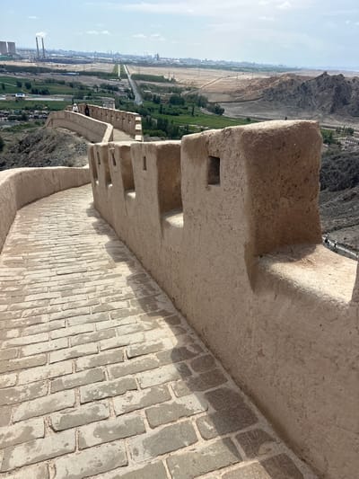The image depicts a section of the Jiayuguan Great Wall in Jiayuguan, China, under a partly cloudy sky. The foreground is dominated by a winding pathway paved with weathered grey bricks, curving to the right. To the right of the path stands a section of the Great Wall constructed from earth and straw, with crenellations and small square openings visible along its top. The wall is a light brown, textured material. In the middle ground, a few individuals are walking along a slightly elevated portion of the wall. Beyond the wall, the landscape transitions to rolling hills and a valley. In the distance, a sprawling industrial area with smokestacks emitting plumes of white smoke is visible, along with a city skyline and power lines stretching across the terrain. The overall scene showcases the juxtaposition of historical architecture with modern industrial development.