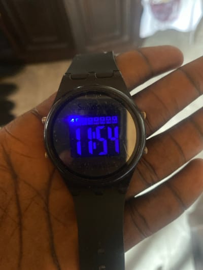 A close-up shot captured in Lakka, Sierra Leone, displays a person's hand holding a black digital wristwatch. The hand, with a dark skin tone, is open, presenting the watch prominently. The watch features a black case and a matching black strap with visible adjustment holes.

The central focus is the watch's vibrant blue LED display, which clearly shows the time as "7:51". To the left of the time, the brand name "Sunkey" is legible in white text. Faintly visible on the right side of the display, the word "WATCH" can be seen. Below the main time display, there are several small blue squares, possibly indicating days of the week or a date, though specific characters are not decipherable. A small, glowing blue icon, possibly representing an alarm or mode indicator, is active near the bottom left of the screen.

The watch face shows some signs of wear, with light scratches or dust visible on its surface. In the background, elements are blurred, suggesting an indoor setting. These indistinct forms include what appears to be a light-colored surface or cloth on the left and a darker, possibly wooden, surface or furniture piece on the right. The overall lighting seems moderate, with the watch's display providing its own illumination. The activity depicted is simply the presentation or wearing of the watch.