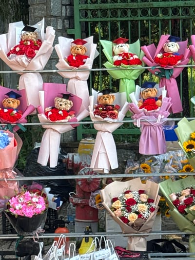 The image depicts a stall in Lâm Viên - Đà Lạt, Vietnam, selling graduation bouquets. The stall features several shelves filled with flower arrangements and teddy bears dressed in graduation attire. In the foreground, on the bottom shelf, are bouquets of pink and white flowers. The middle shelves showcase more elaborate bouquets, some containing teddy bears wearing graduation caps and gowns, accompanied by red roses. The background reveals a green metal fence and a stone wall. The overall scene suggests a festive atmosphere, likely a popular spot for students celebrating graduation.