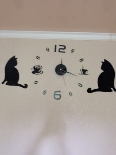 The image shows a wall clock with a black cat motif. The clock has large, black Arabic numerals for 12 and 6, and smaller coffee bean shapes serving as hour markers. Two black silhouette cats are positioned on the left and right sides of the clock face. A coffee cup silhouette is also present. The clock hands indicate a time slightly before 10:15. The wall behind the clock appears to be a pale, textured surface, possibly wallpaper or painted plaster. The top of the image shows a white architectural trim, likely a crown molding. The lighting suggests the image was taken indoors, possibly in a residential or commercial setting. There are no people or any other distinguishing environmental features visible that would allow for a specific location within Temirtau, Kazakhstan to be identified.