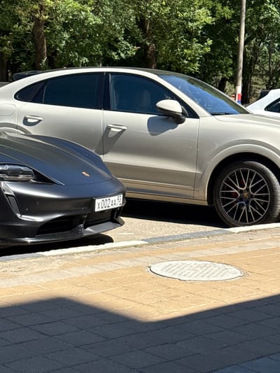 Two Porsche vehicles are parked on a street in Armavir, Russia. The vehicle in the foreground is a dark gray electric Porsche Taycan, and its license plate reads "X002AA 93". The vehicle behind it is a light-colored Porsche Cayenne. The cars are parked on a paved street next to a sidewalk made of yellow and gray rectangular pavers. A circular manhole cover is visible on the sidewalk. Trees with green foliage line the background, and a lamppost is visible behind the Cayenne. The scene appears to be a daytime shot, with the sun casting shadows on the ground.