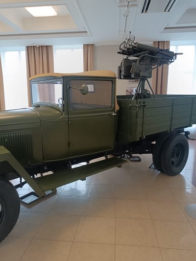 The image shows a vintage military truck with a mounted machine gun, displayed indoors. The truck is olive green, with a canvas roof over the cabin and wooden sides on the cargo bed. The machine gun is positioned on a tripod mount in the rear of the truck bed. The setting appears to be a museum, with light-colored tiled flooring and large windows with beige curtains in the background. Above the truck, a modern ceiling with recessed lighting fixtures is visible. The environment is clean and well-lit. This artifact is part of the military technology exhibition at the Museum of Military Technology "Battle Glory of the Urals" in Verkhnyaya Pyshma, Russia. No people are present in the image.