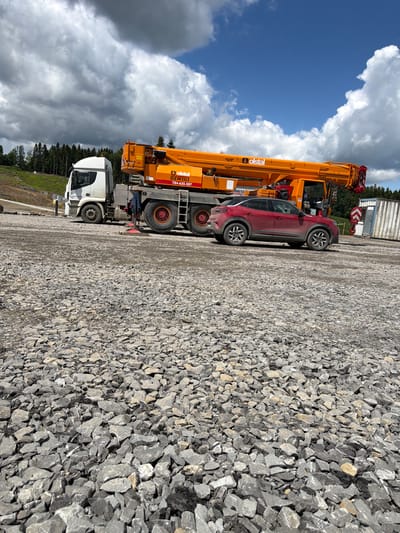 The image depicts a construction site in Skrzydlna, Poland, characterized by a vast expanse of crushed grey stone gravel covering the ground. Dominating the scene is a large orange crane truck with a white cab, parked on the gravel. Next to the crane truck, a red SUV is also parked. In the background, a line of dark green pine trees forms the horizon, with a blue sky with scattered white clouds above. A white shipping container with a red hook attached to it is visible to the right of the crane truck. The overall impression is one of an active outdoor construction or industrial area.