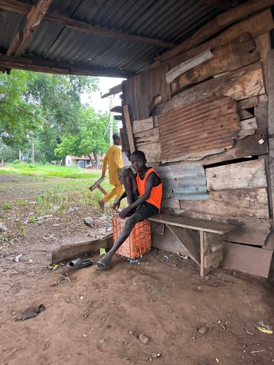 This image depicts an outdoor scene in Idah, Nigeria, featuring a makeshift structure and two males.
In the foreground and to the right, a rudimentary shelter is constructed from rough-hewn wooden planks and corrugated metal sheets. The roof, made of corrugated metal, is supported by visible wooden beams. The right wall of the structure is assembled from various weathered wooden boards and rusted corrugated metal panels. A small, unpainted wooden bench or shelf is affixed to this wall at a low height. A light-colored padlock is visible on a wooden plank in the upper right section of the wall.
A young male, appearing to be a child or adolescent, is seated on an orange plastic crate on the left side of the shelter. He wears an orange sleeveless top and dark shorts. His gaze is directed towards the viewer. His left leg is extended forward, and his right leg is bent. A pair of black slide sandals are on the reddish-brown earthen ground beside his feet; one is off his foot, and the other is partially on his right foot.
In the mid-ground, another male walks from left to right. He is dressed in a loose-fitting, long-sleeved, light yellow or mustard-colored garment. He carries a dark, angular wooden object, possibly a small frame or stool, in his right hand. His face is turned away from the camera.
The ground throughout the scene is composed of reddish-brown soil, interspersed with sparse patches of green vegetation and scattered debris, including plastic wrappers and discarded items. In the mid-ground and background to the left, an open, unkempt grassy area with numerous green-leafed trees is visible. Utility poles and overhead lines are present in the background. Further back, several simple structures with corrugated metal roofs are discernible, with one building featuring a blue tank or container beside it.