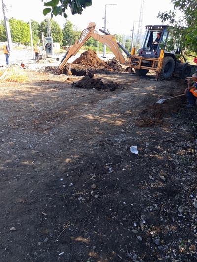 A construction site is active in Dragoman, Bulgaria, with two excavators and several workers present. In the foreground, a man wearing an orange vest sits on the ground, holding a shovel. Behind him, another man in a similar vest operates a backhoe loader. To the left, a third worker in a hard hat and vest stands near a smaller excavator. The ground is uneven and covered with dirt, rocks, and construction debris. Piles of excavated earth are visible, and a partially constructed trench runs through the site. Power lines and trees are visible in the background.