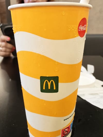 A close-up, eye-level shot shows a cold, condensation-covered disposable cup with white and orange horizontal stripes. A red circular Coca-Cola logo is visible on the upper right side of the cup, and a green square with the McDonald's golden arches logo is on the lower center. The cup is sitting on a dark, reflective surface, likely a table. In the background, a person's hands are visible, holding a smartphone. The person has painted fingernails. The overall scene suggests a fast-food setting, possibly a McDonald's in Naples, Italy, given the presence of the McDonald's and Coca-Cola branding.