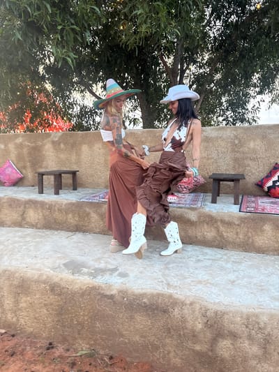 Two women are standing on a stepped stone structure in an outdoor setting, possibly a resort in Mui Ne, Vietnam. The woman on the left wears a white bandeau top, a long brown skirt, and a colorful wide-brimmed hat. She has tattoos on her arms and chest. The woman on the right wears a white and brown cow-print halter top, a brown ruffled skirt, and a white cowboy hat. She also has tattoos. Both women are wearing white cowboy boots. They are facing each other, with their hands intertwined, and appear to be interacting playfully. The environment is rustic, with natural stone steps, some decorative pillows, and small wooden stools. Green foliage from a tree is visible in the background, along with warm lighting that suggests it might be late afternoon or early evening.