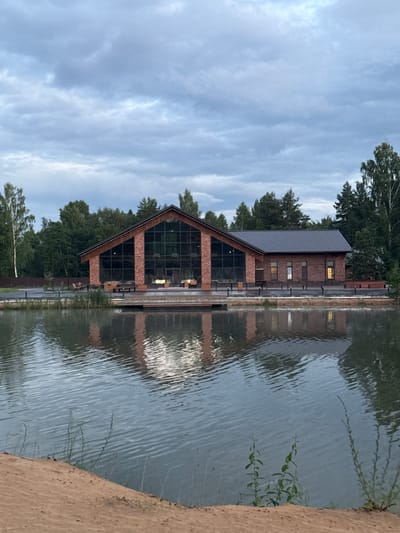A contemporary building with a pitched, dark-colored roof and brick walls stands on the edge of a body of water. The building features a large, triangular section with floor-to-ceiling windows, reflecting the sky and surrounding trees. Other windows are visible higher up on the brick facade. The water's surface is rippled, creating a distorted reflection of the building and the trees. The sky is overcast with light, wispy clouds. Green trees line the background behind the building, and some foliage is visible on the right side of the image, extending into the water. A sandy or dirt embankment is visible in the foreground on the right. The scene appears to be set in a natural environment, possibly a park or recreational area, in Sosnovka, Russia. The time of day seems to be either early morning or late evening, given the muted light and overcast conditions. No people or text are visible in the image.