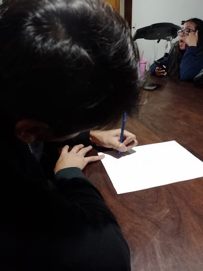 The image depicts a dimly lit indoor scene, likely in a home in Gobernador Julio A. Costa, Argentina, given the context provided. A person with dark hair is seated at a dark wooden table, focused on writing on a white sheet of paper with a blue pen. The person is wearing a dark sweatshirt. In the background, another person with glasses is visible, seated at the same table and holding a smartphone. A pink cup, another smartphone, and a piece of exercise equipment are also visible on the table in the background. The primary action is the act of writing.