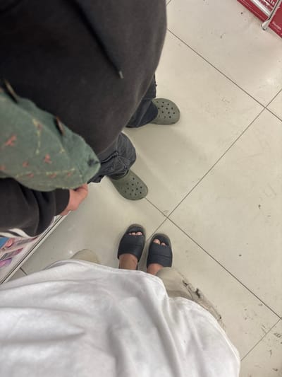 The image depicts the lower extremities of two individuals standing on a light-colored tiled floor within an indoor commercial or retail establishment in Pai, Thailand.
In the foreground, a person is visible from the waist down, wearing a flowing white garment, possibly a skirt or robe, and dark blue or black slide sandals. Their bare feet are partially visible within the sandals.
In the mid-ground, a second individual stands directly in front of and slightly to the right of the foreground person. This individual wears dark grey or black trousers, an olive-green hooded sweatshirt, and olive-green Crocs-style clogs. A green fabric bag with a faint pattern of small reddish shapes hangs from their left arm/shoulder area.
The floor is composed of large, light-colored square tiles with dark grout lines, showing minor scuff marks or debris. On the far left, a vertical white shelf or display unit is partially visible. In the upper right corner, a portion of a red object, possibly a metal shelf edge or sign, is discernible. Both individuals appear to be standing still, facing forward relative to the camera's perspective. No direct interaction between the individuals is discernible.