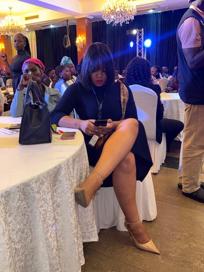 The image depicts an indoor event, likely a conference or formal gathering, at a location in Itirin, Nigeria.

In the foreground, a woman with dark, shoulder-length hair and bangs is seated at a white-draped table, engrossed in a black smartphone held in both hands. She wears a black long-sleeved dress, nude-colored pointed-toe high heels with ankle straps, and an event lanyard around her neck. Her legs are crossed, and a patterned scarf is draped over her right shoulder. On the table to her left, a black handbag, a pink smartphone, and some papers are visible.

To the immediate left of the main subject, an older woman with a pink headscarf and a light blue jacket is seated, looking to her right. A black handbag rests on the table in front of her. Further back and to the left, another woman in a black turtleneck stands or leans, facing right. Numerous other attendees are visible in the mid-ground, mostly seated, with a woman in a light blue headwrap partially discernible. To the right of the main subject, the lower torso and legs of a standing man are visible, wearing a light-colored vest, pink shirt, tan trousers, and dark slip-on shoes.

The background reveals a large room or ballroom featuring ornate chandeliers with warm lighting. A dark blue or black curtain serves as a backdrop, illuminated by blue stage lights, and a metal truss structure is visible. The floor is tiled.