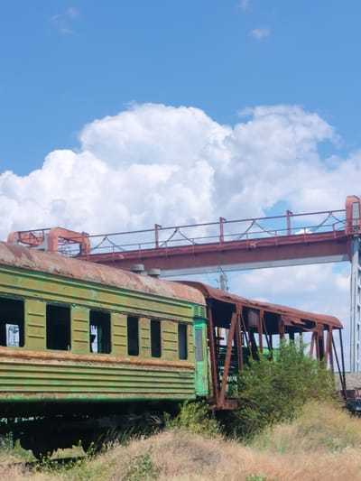 The image depicts an abandoned train yard under a bright blue sky with puffy white clouds. In the foreground, two derelict train cars sit on tracks, partially obscured by dry grass and weeds. The first car, painted green, shows significant rust and peeling paint, with its windows black and empty. To its right is a rusted metal structure, possibly the remains of another train car or a bridge. In the background, a large, rusty industrial gantry or bridge structure spans across the scene, supported by tall concrete pillars. The overall impression is one of decay and disuse. The specific location in the city of Salsk, Russia, is not visually indicated by any identifiable landmarks within the image.